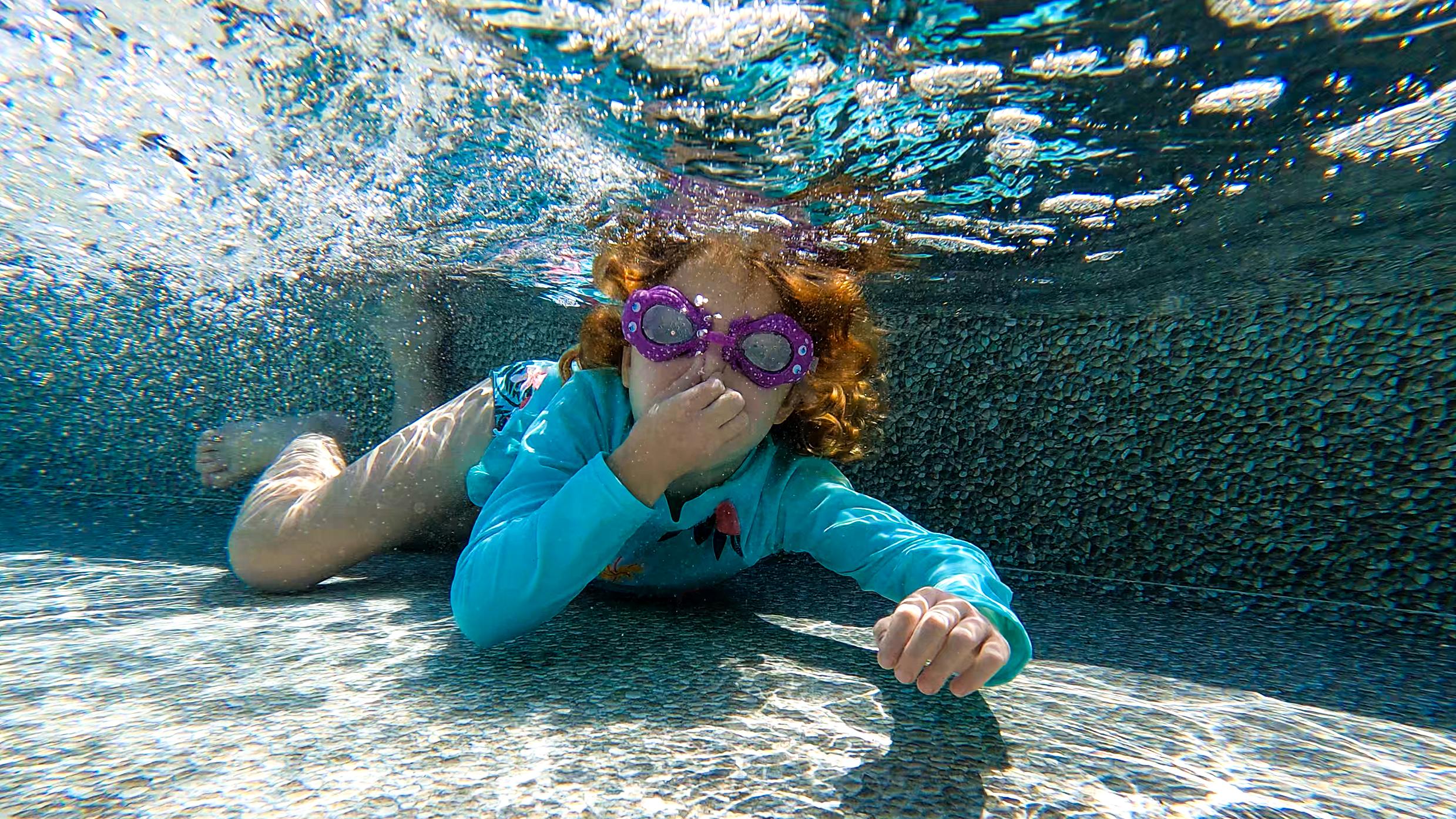 Girl in purple goggles holding her nose underwater