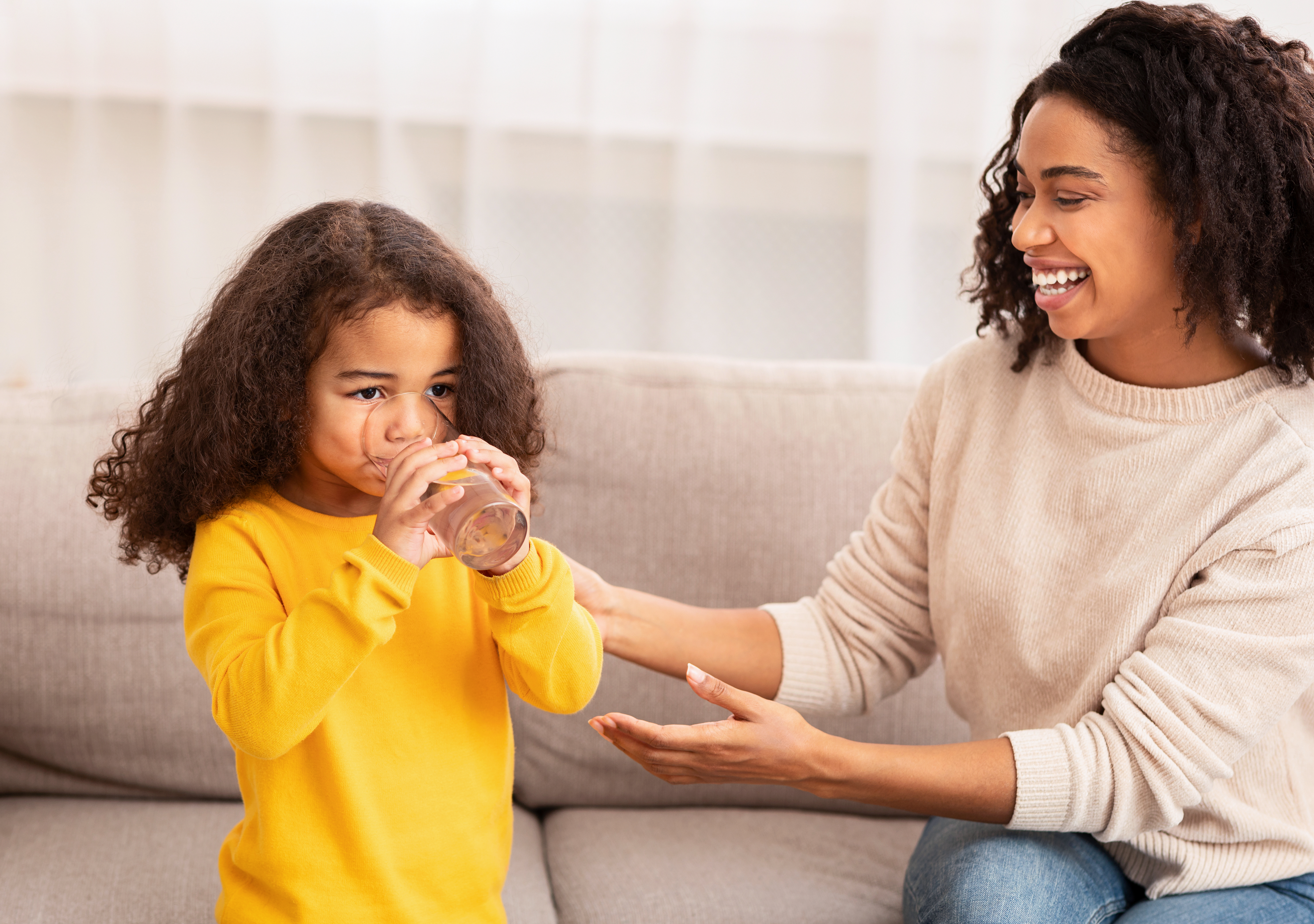 Hero Image Mother's Care. Joyful African American Woman Feeding Little Daughter Giving Her Glass Of Water Sitting On Sofa At Home.; Gettyimages: 1199674829