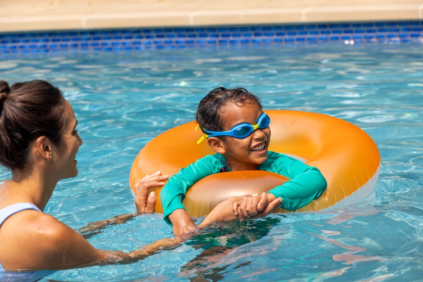 little boy in pool wearing swimming goggles and a tube floaty with mom