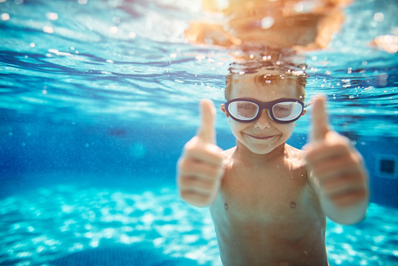 Little boy aged 6 swimming underwater. The boy is smiling at the camera showing thumbs up.