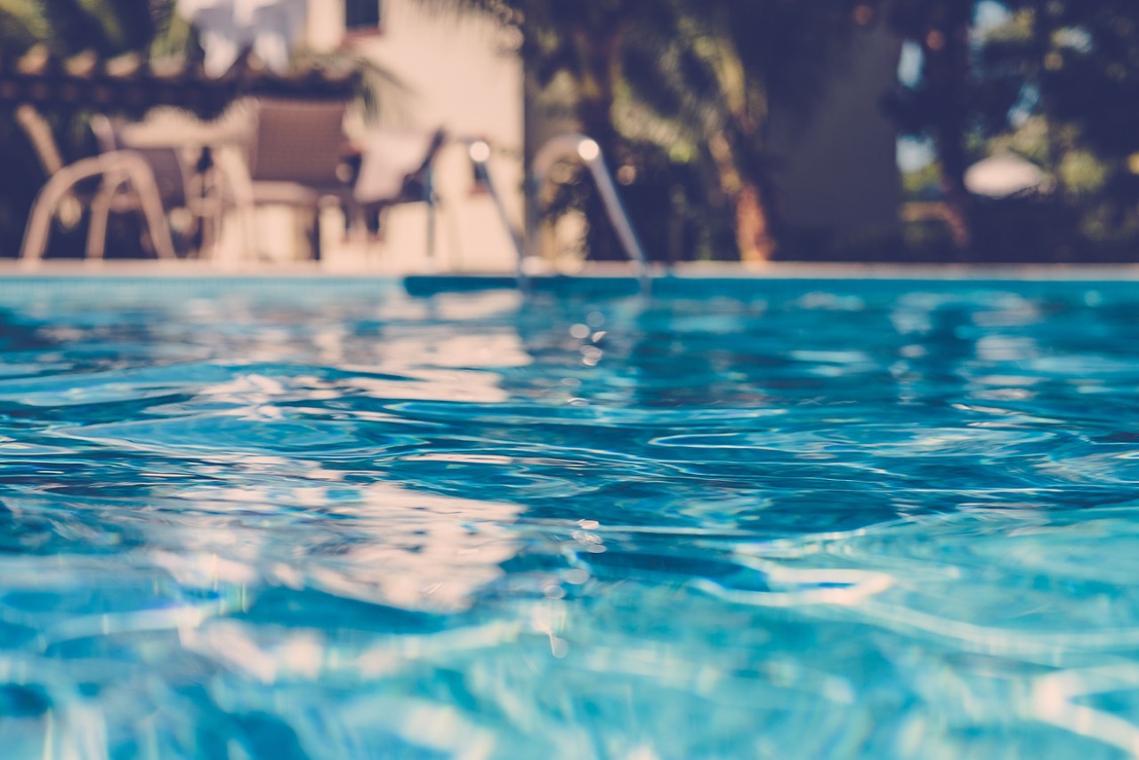 blurred surface view on blue swimming pool water with trees and table and chairs in the background