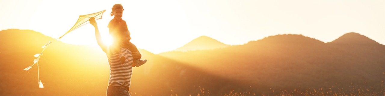 Adult male with little girl on shoulders flying kite in field during golden hour