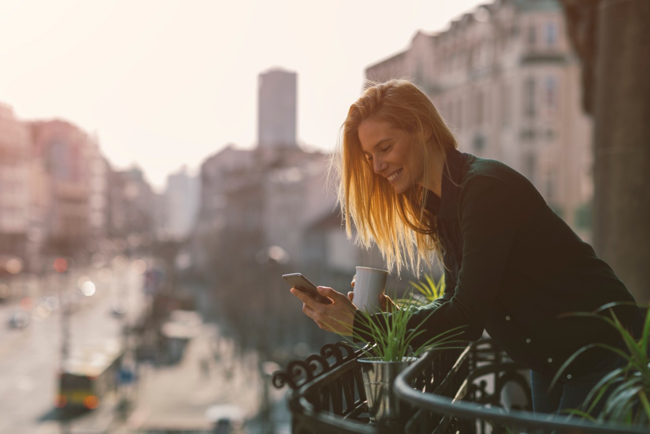 Single woman drinking coffee and using her smart phone on hotel balcony