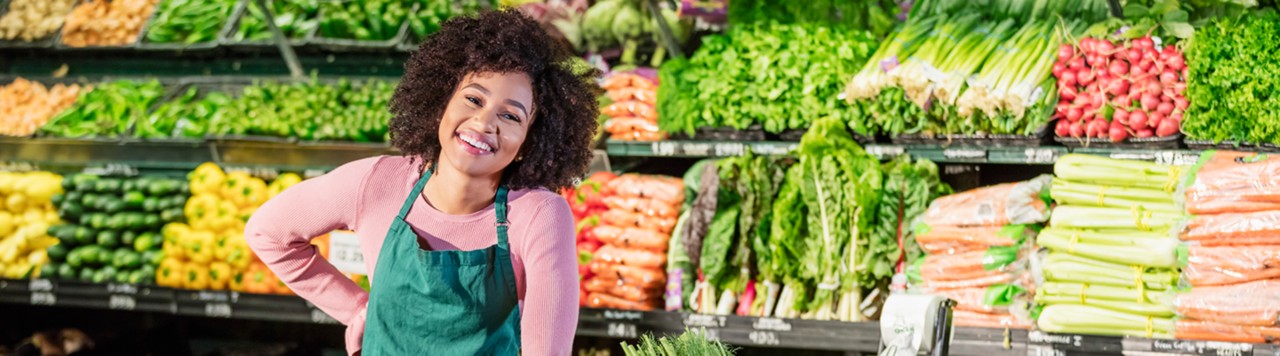 A young African-American woman in her 20s working in the produce aisle of a grocery store, surround by fresh fruits and vegetables.