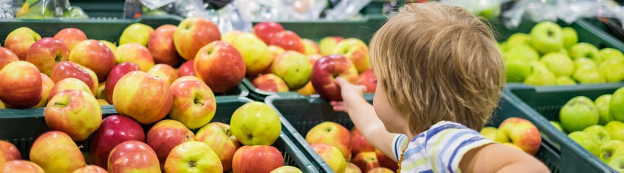 A young boy in the produce aisle of a grocery store, reaching for an apple.; Gettyimages: 960638210