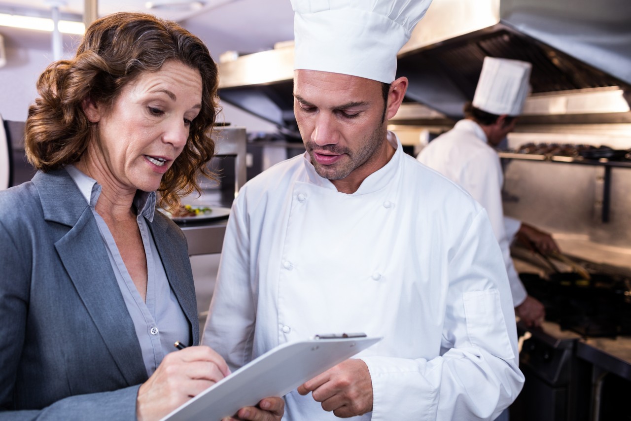 Female restaurant manager writing on clipboard while interacting to head chef in commercial kitchen; Shutterstock ID 564107209; Project: TWM WEB PAGE