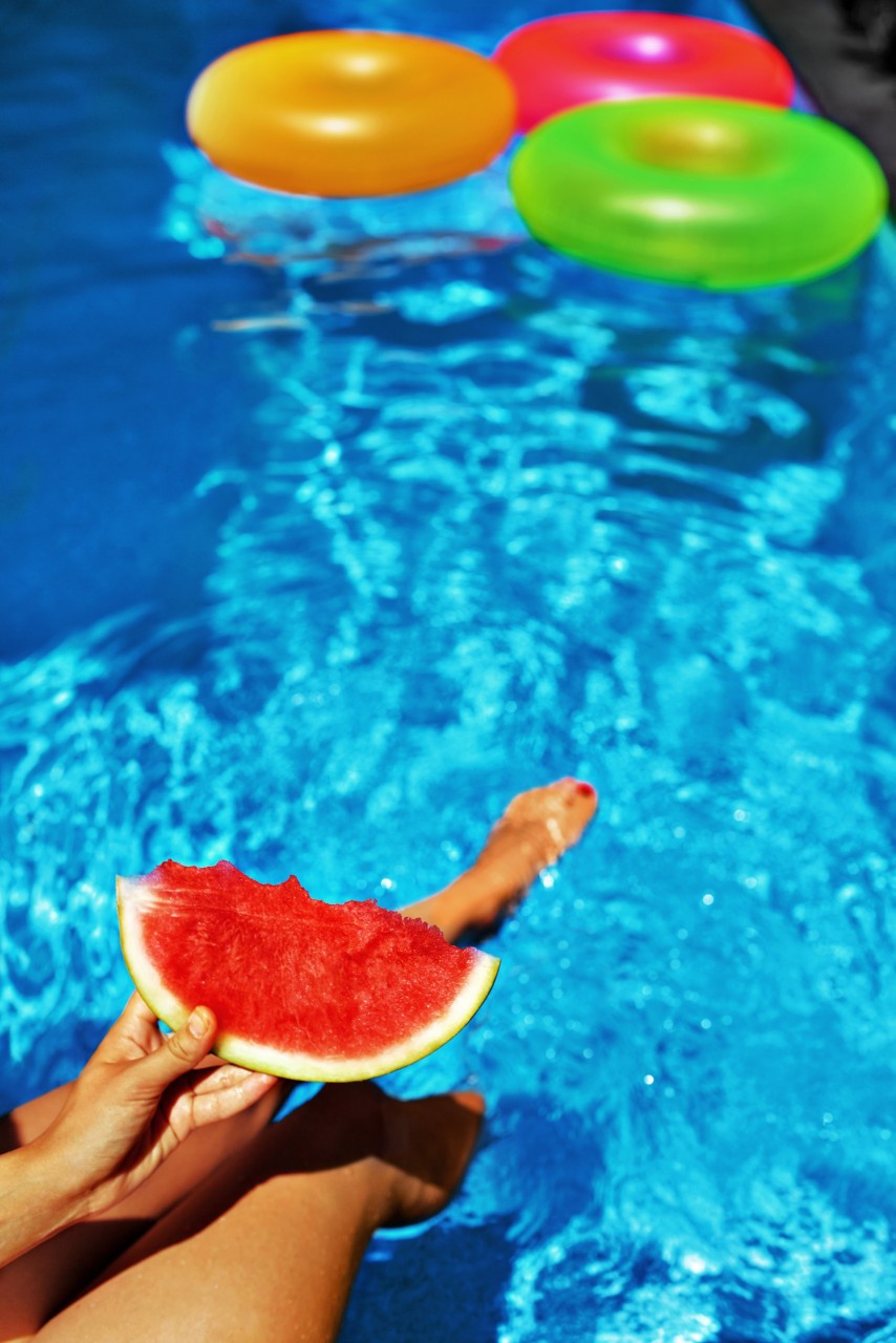 Female Hand Holding Slice Of Ripe Juicy Watermelon By The Swimming Pool. Colorful inflatable Floating tubes In Refreshing Blue Water On Background