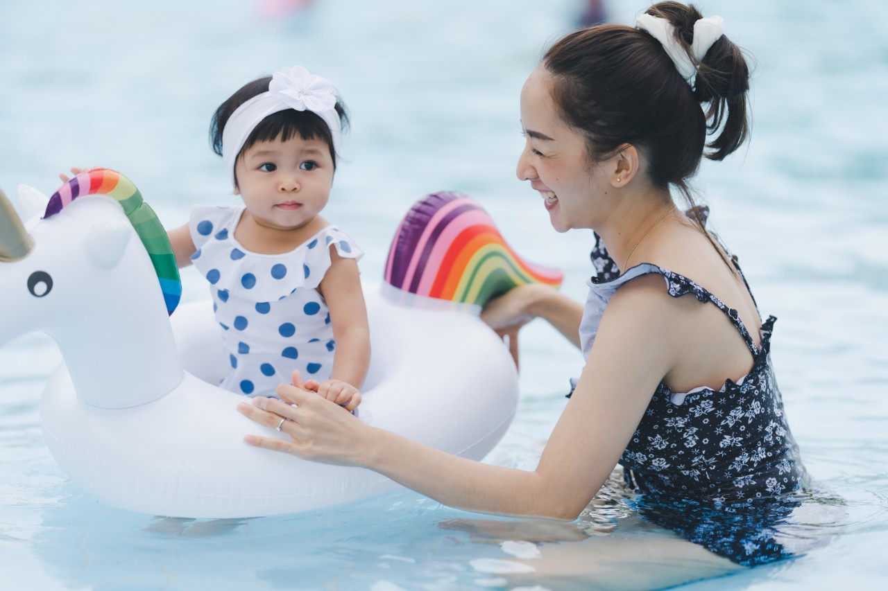 An asian mother and child are playing in shallow water. The young girl is in a rainbow unicorn float