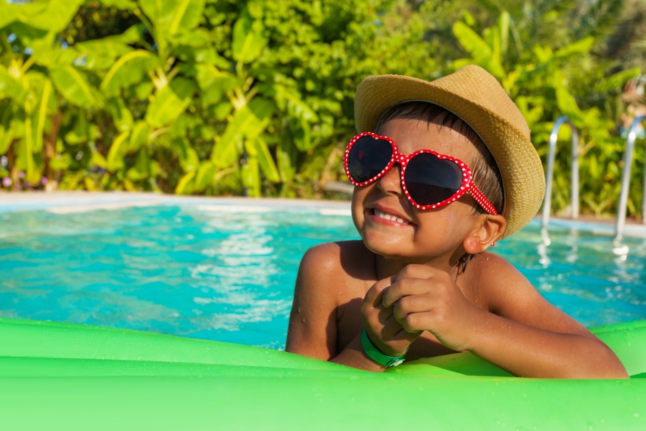 Cute boy in heart-shaped sunglasses and hat on green airbed inflatable in the swimming pool outside in summer