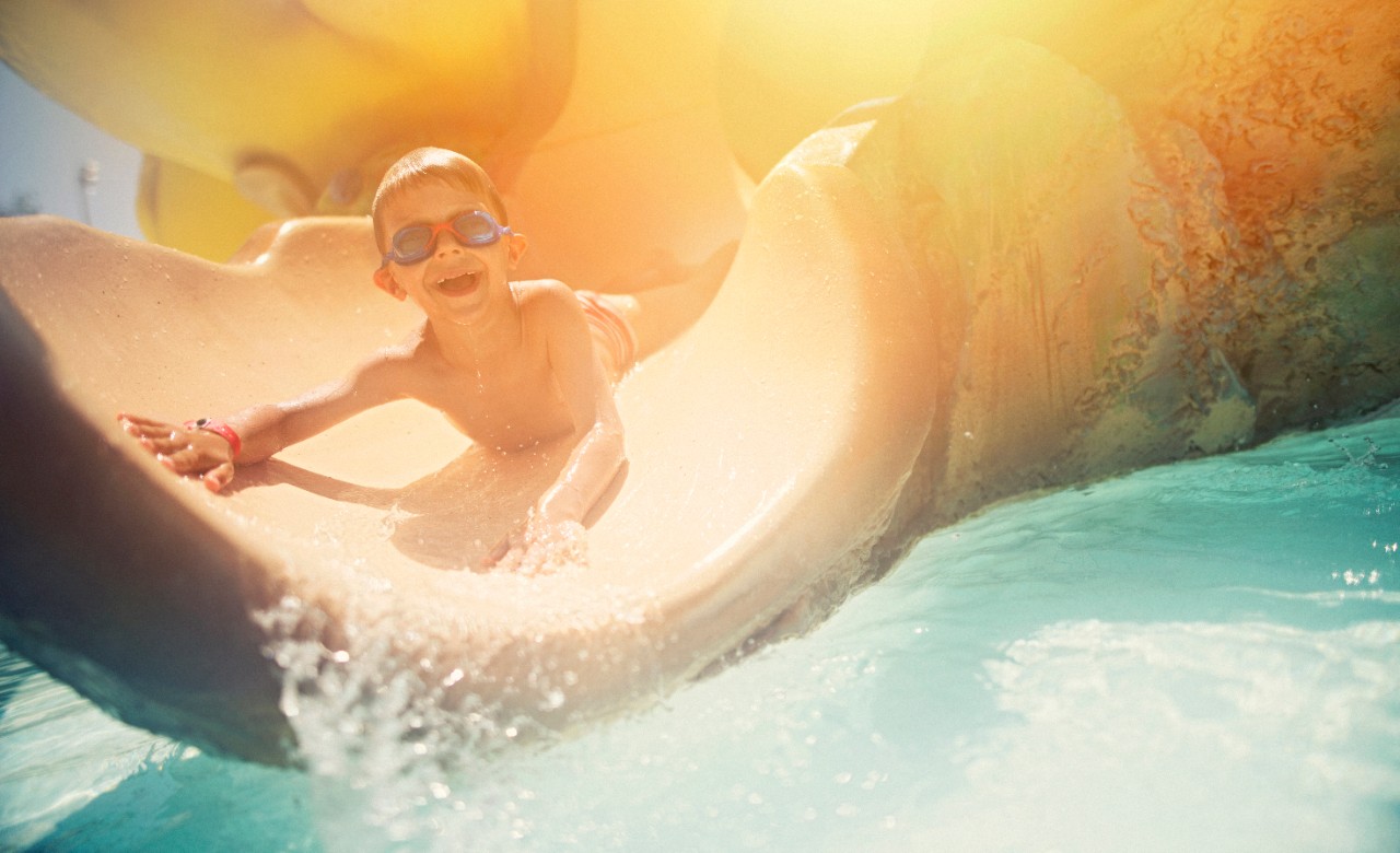 Little boy sliding on water slide. The funny boy aged 5 is wearing swimming goggles. Sunny summer day on mallorca.