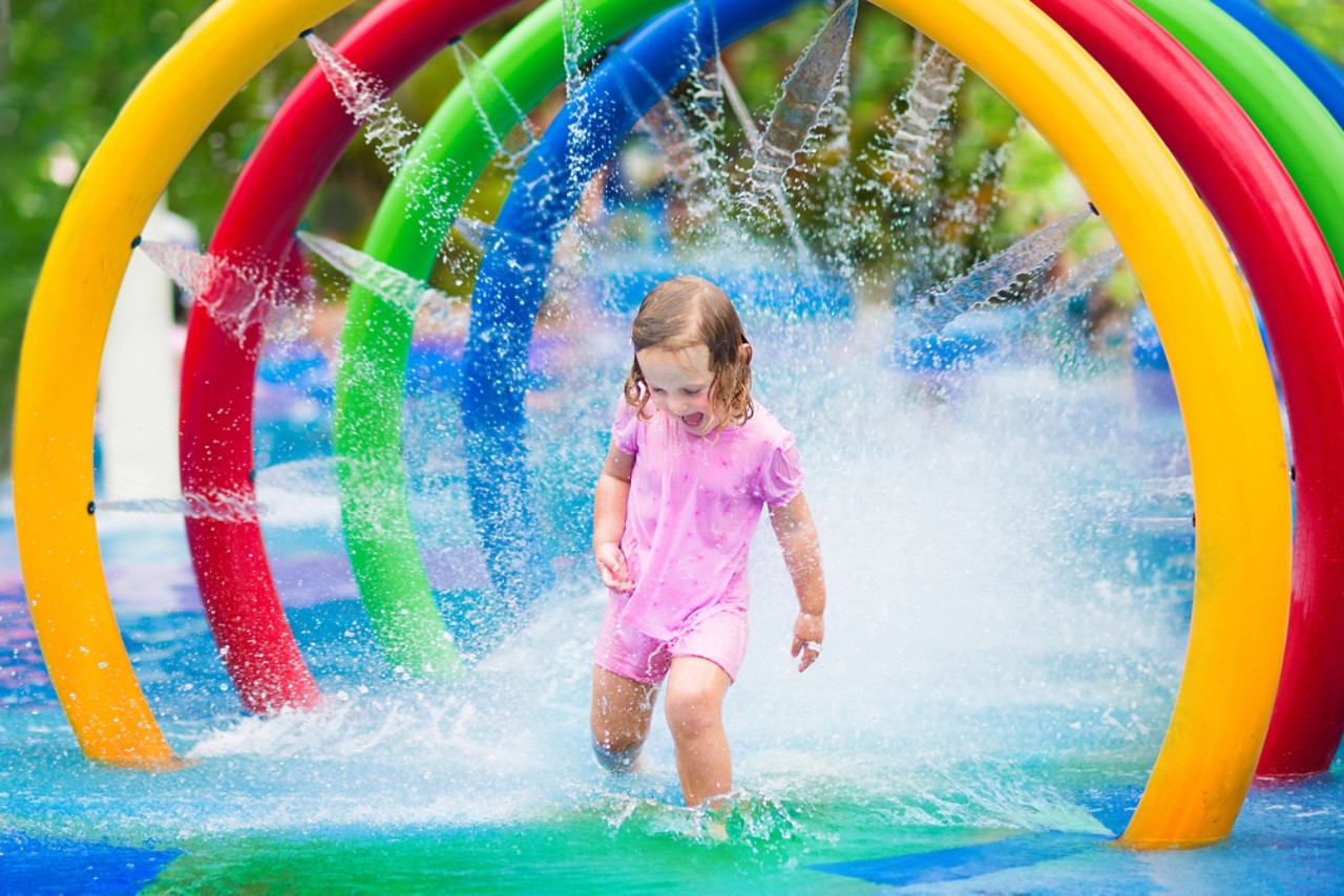 Happy little toddler girl running through a inflatable toy sprinkler having fun with water splashes in a swimming pool enjoying day trip to an aqua amusement park during summer family vacation