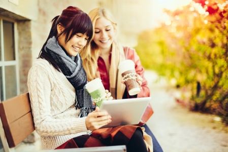 young-women-looking-at-tablet-while-drinking-coffee-on-outdoor-bench-in-fall-with-colorful-leaves-horizontal-5000x3333-image-file-496515403