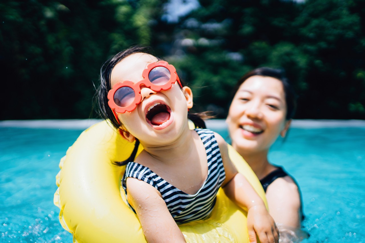 Happy toddler girl with sunglasses smiling joyfully and enjoying family bonding time with mother having fun in the swimming pool in summer