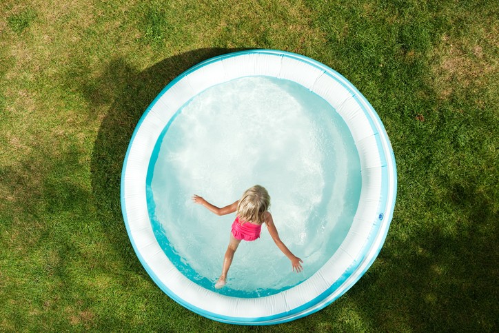 A six year old girl is jumping in an inflatable swimming pool on a hot Summer day. The grass is becoming dry and brown.