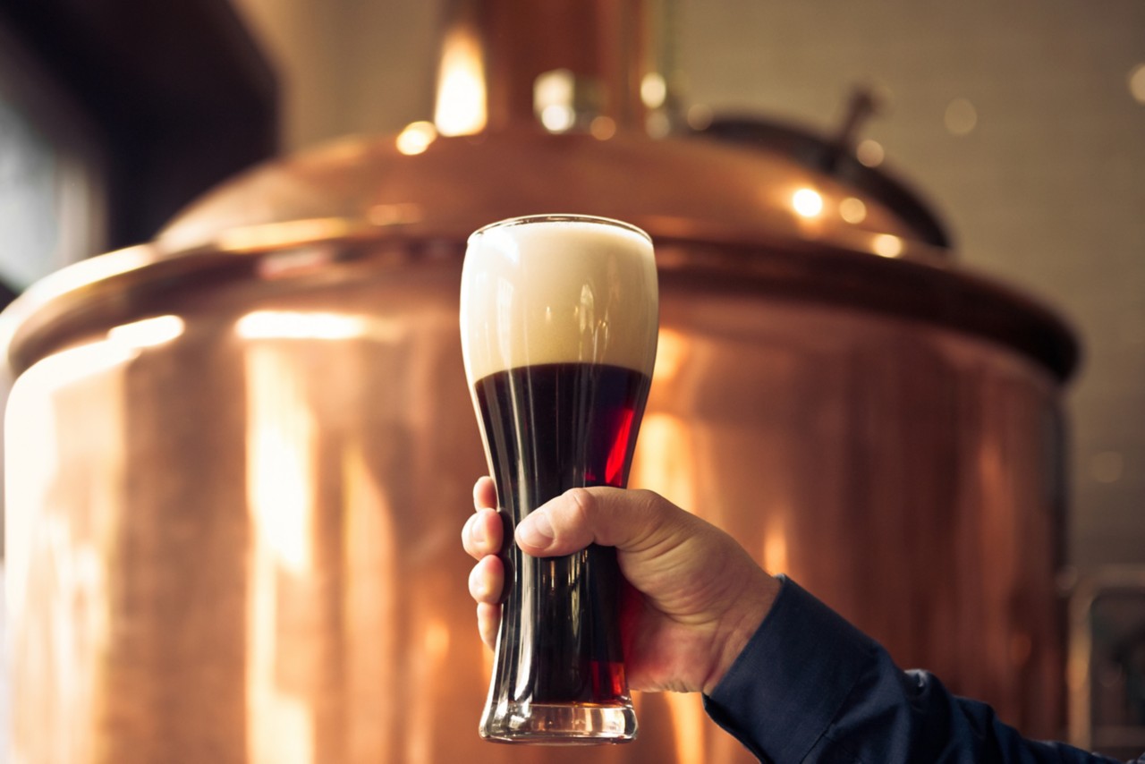 Close up of male brewer holding glass of bitter ale beer in front of copper vat