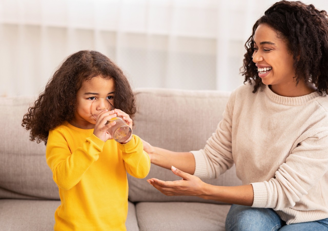 Mother's Care. Joyful African American Woman Feeding Little Daughter Giving Her Glass Of Water Sitting On Sofa At Home.