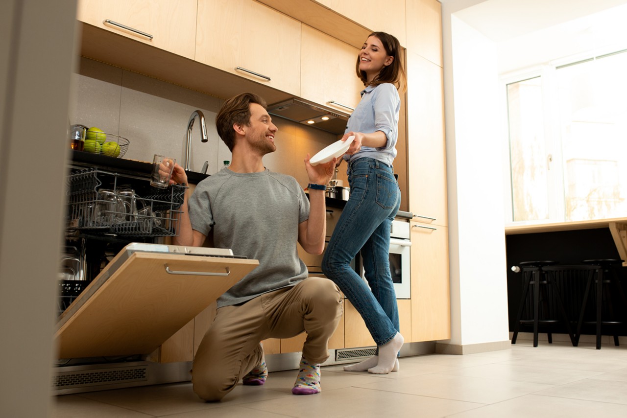 Smiling male is putting dishes into dishwasher with pretty lady on the background at home; Shutterstock ID 1733843165; purchase_order: pws website; job: 