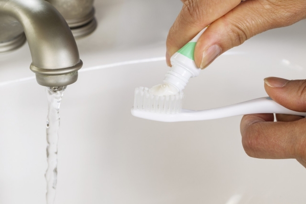 female hands putting tooth paste on toothbrush with white bathroom sink and running faucet in background 