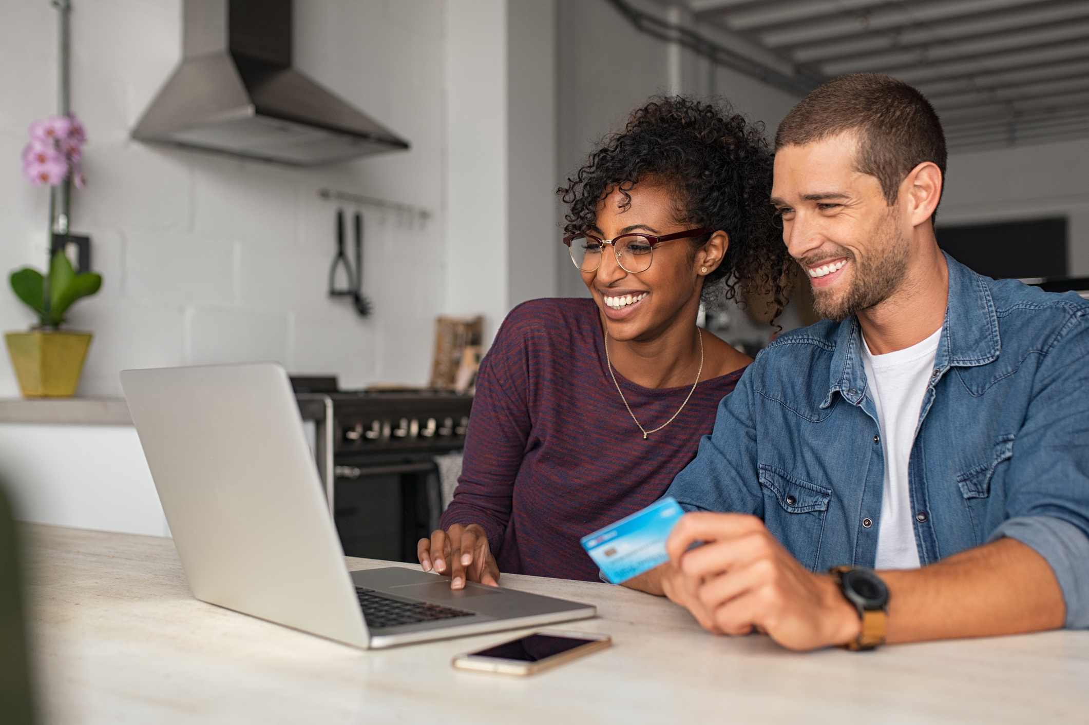 Smiling young couple making shopping online with credit card and laptop at home. Happy multiethnic couple holding debit card while buying on ecommerce site using laptop. Cheerful guy and african girl making online purchase.